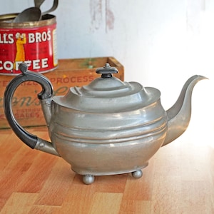 May include: A vintage silver teapot with a black handle and a spout. The teapot sits on a wooden surface, with a can of Hills Bros. Coffee and a wooden box in the background. The teapot has a rounded body and three small feet.