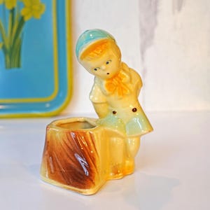 May include: A ceramic planter shaped like a girl wearing a blue hat and a green and blue dress. The planter is standing on a brown tree stump.