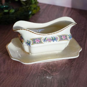 May include: A white porcelain gravy boat with a floral and bird design. The boat has a gold trim and is sitting on a matching saucer.