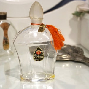 May include: A clear glass bottle with a frosted glass stopper and an orange tassel. The bottle has a label that reads "Ex-Celcis".