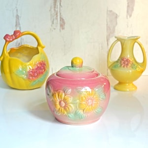 May include: Three ceramic decorative pieces: a yellow basket with a pink bow, a pink lidded jar with yellow flowers, and a yellow vase with handles. All pieces have floral designs and are on a white surface.