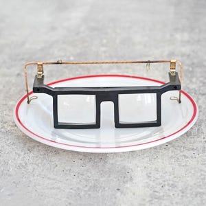 May include: A pair of black-framed glasses with clear lenses rests on a white plate with a red rim. A copper wire and gold-colored metal pieces are attached to the glasses. The plate sits on a gray surface.