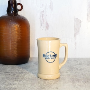May include: A tall, cream-colored ceramic mug with a handle, featuring the blue text Buckeye Root Beer inside a circular emblem. Beside it stands a large, dark amber glass jug with a handle, embossed with the words One Gallon.