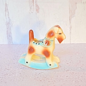 May include: A vintage ceramic rocking dog figurine. The dog is cream-colored with brown spots and a brown tail. It has a light blue saddle and is mounted on a light blue rocking base. The figurine is a decorative item.