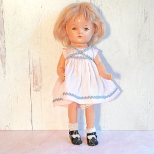 May include: A vintage doll with blonde hair, wearing a pink dress with blue trim and black shoes.