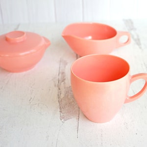 May include: A set of three pink plastic dishes, including a small bowl with a lid, a creamer, and a mug.