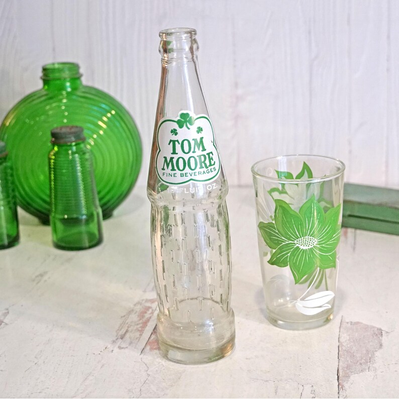 May include: A clear glass Tom Moore Fine Beverages bottle with a green and white label, and a clear glass with a green floral design. Several green glass bottles are in the background.