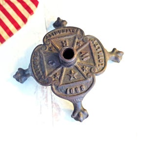 May include: A cast iron decorative stand with a cross-shaped design. The stand has four ornate feet and a central hole. The words "AMITY", "LOYALTY", and "1888" are visible. A small American flag is in the background.