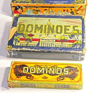 May include: Three vintage domino game boxes. The top box is red, white, and blue with the text "Toys That Teach" and "The Embossing Company Made in USA". The middle box is blue and white with the text "Dominoes" and "930". The bottom box is yellow and green with the text "Dominoes" and a dragon design.