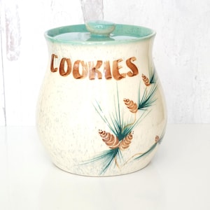 May include: Vintage ceramic cookie jar with a cream-colored base and a turquoise rim and lid. The word "COOKIES" is written in brown script. The jar is decorated with green and brown pine needle and cone designs.