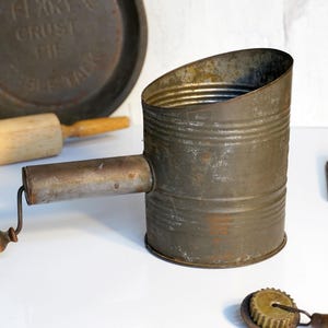May include: Vintage metal flour sifter with a wooden handle and a crank. The sifter is rusty and has a worn finish.