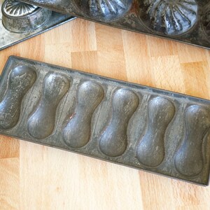 May include: A vintage metal baking pan with eight individual molds shaped like elongated ovals. The pan is made of a dark gray metal and has a worn, rustic appearance.