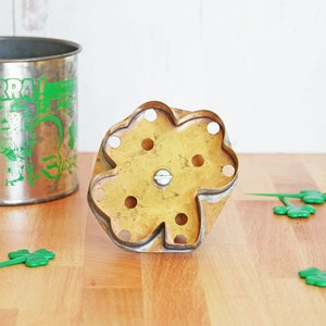May include: A vintage metal cookie cutter shaped like a four-leaf clover. The cutter is brass colored and has eight holes around the edges.
