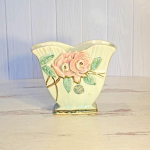 May include: A vintage ceramic vase with a light green base and a scalloped top. The vase features a raised design of pink roses with green leaves and gold accents. A small, round emblem is visible on the front.
