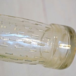 May include: A clear glass bottle with a textured, rectangular pattern. The bottle has a cylindrical shape with a rounded base and a slightly wider neck. The glass has a slight amber tint, and the bottle is set against a light background.