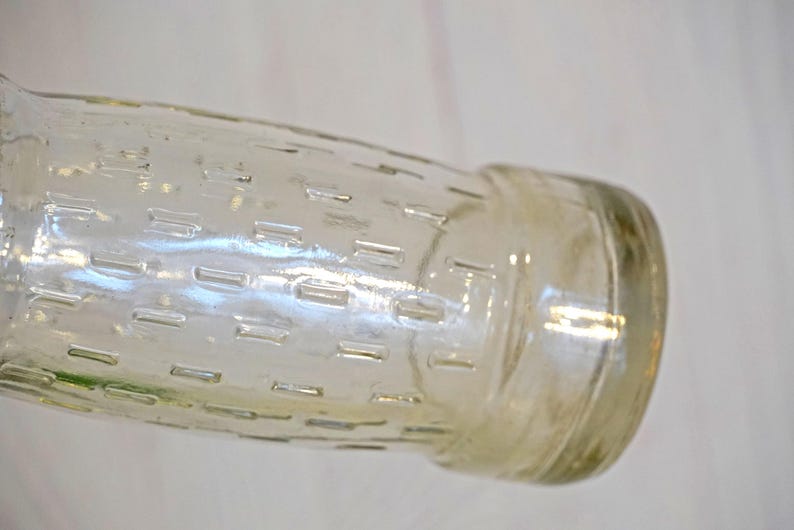 May include: A clear glass bottle with a textured, rectangular pattern. The bottle has a cylindrical shape with a rounded base and a slightly wider neck. The glass has a slight amber tint, and the bottle is set against a light background.