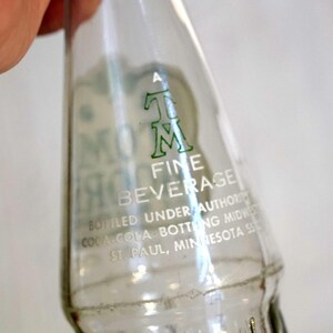 May include: Clear glass bottle with green lettering. The bottle reads "MOM FINE BEVERAGE" and "Bottled Under Authority Coca Cola Bottling Midwest St. Paul, Minnesota". The bottle is held by a person.