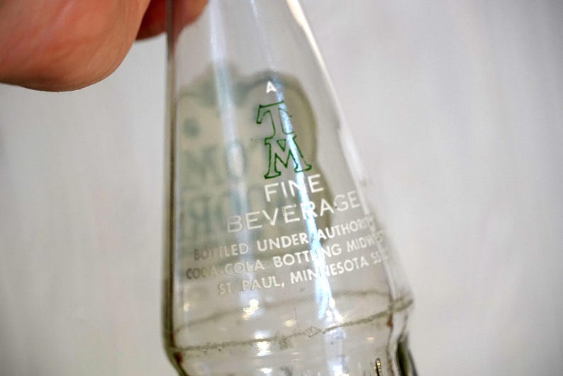 May include: Clear glass bottle with green lettering. The bottle reads "MOM FINE BEVERAGE" and "Bottled Under Authority Coca Cola Bottling Midwest St. Paul, Minnesota". The bottle is held by a person.