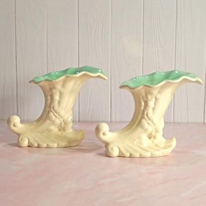 May include: Two vintage ceramic vases. The vases are a creamy yellow with a light green ruffled rim. The vases have a curved shape with a decorative scroll base and embossed floral details. They are displayed on a pink surface.