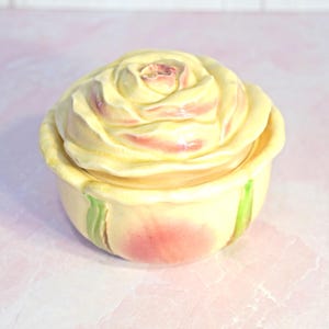 May include: A ceramic rose-shaped trinket box. The lid and base are a pale yellow with pink and green accents. The box is designed to resemble a blooming rose, with detailed petal formations.