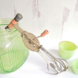 May include: A vintage hand-crank egg beater with a green glass jar and two green glass measuring cups. The egg beater has a metal crank and a set of metal beaters.