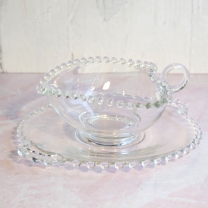 May include: A clear glass gravy boat with a beaded rim and handle, sitting on a matching saucer.