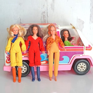 May include: A vintage toy set featuring a pink and white van with a colorful design, accompanied by three dolls in brightly colored jumpsuits. One doll is in the driver's seat. The dolls are wearing yellow, red, and orange outfits.
