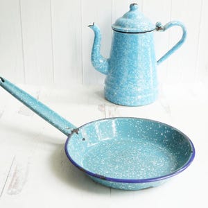 May include: A blue enamel frying pan with a speckled finish and a blue rim. The pan has a long handle and is sitting on a white surface.