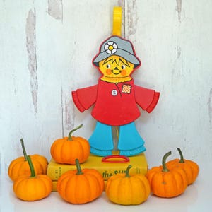 May include: A plastic scarecrow toy with a red shirt, blue pants, and a gray hat with a flower. The scarecrow is standing on a stack of books with the title "The Sun" visible. There are several small orange pumpkins in front of the scarecrow.
