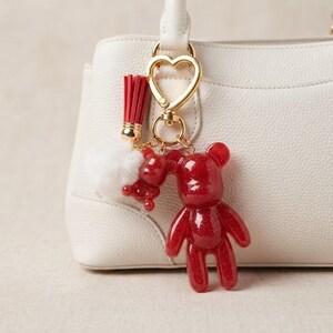 May include: A white textured handbag adorned with a gold heart-shaped keychain. The keychain features a red glittery bear charm, a smaller matching red bear, a red tassel, and a fluffy white pom-pom attached to the gold metal hardware.
