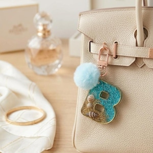 Op de afbeelding: Een sleutelhanger in de vorm van de letter B met een harsontwerp met blauwe glitters aan de bovenkant, gouden glitters aan de onderkant en ingebedde schelpen. De hanger bevat een lichtblauwe pluizige pompon en een roségouden karabijnhaak.