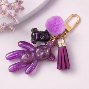 Puede incluir: Un llavero con un dije de oso grande de color morado traslúcido con purpurina, un dije de oso más pequeño de color morado oscuro, un pompón morado esponjoso y una borla de ante morado oscuro unidos a un cierre de mosquetón dorado.