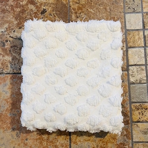 May include: A square, white bath mat with a textured, tufted pattern. The mat has a soft, plush appearance and a fringed edge. The design features evenly spaced, raised circular tufts across the surface.
