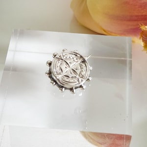 May include: A small, silver-toned brooch with a detailed compass rose design. The brooch is round with a textured border and is displayed on a clear acrylic block. The background includes a white surface and a tulip with yellow and pink petals.