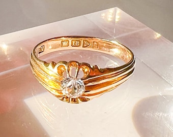 May include: A gold ring with a single diamond set in a raised, ornate setting. The ring is marked with the inscription "18 CT" on the inside band.