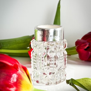 May include: A clear, cut-glass vase with a silver rim. The vase has a geometric pattern and is surrounded by red and green floral elements. The silver rim has markings. The vase is likely for decorative purposes.