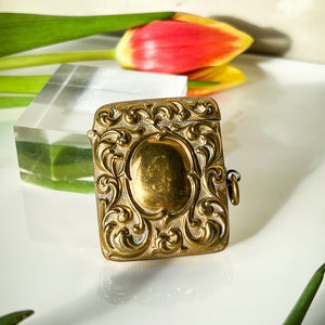 K&ouml;nnte beinhalten: Eine verzierte, rechteckige Schmuckschatulle in Gold, mit einem Scharnierdeckel und einer kleinen Kette. Die Schachtel hat ein detailliertes Blumenmuster und eine glatte, ovale Mitte.