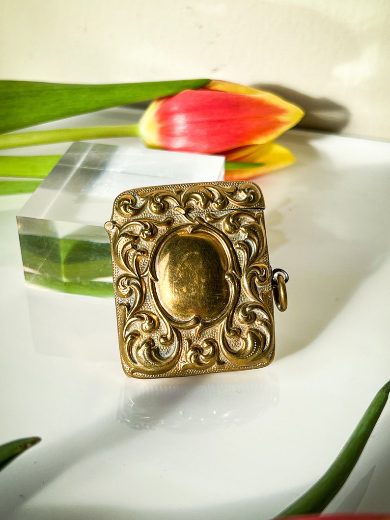 K&ouml;nnte beinhalten: Eine verzierte, rechteckige Schmuckschatulle in Gold, mit einem Scharnierdeckel und einer kleinen Kette. Die Schachtel hat ein detailliertes Blumenmuster und eine glatte, ovale Mitte.
