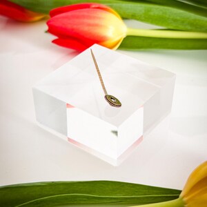 May include: A gold-toned stick pin with a small, round, green and gold decorative element. The pin is displayed on a clear, square block, with red and yellow tulips and green leaves in the background.