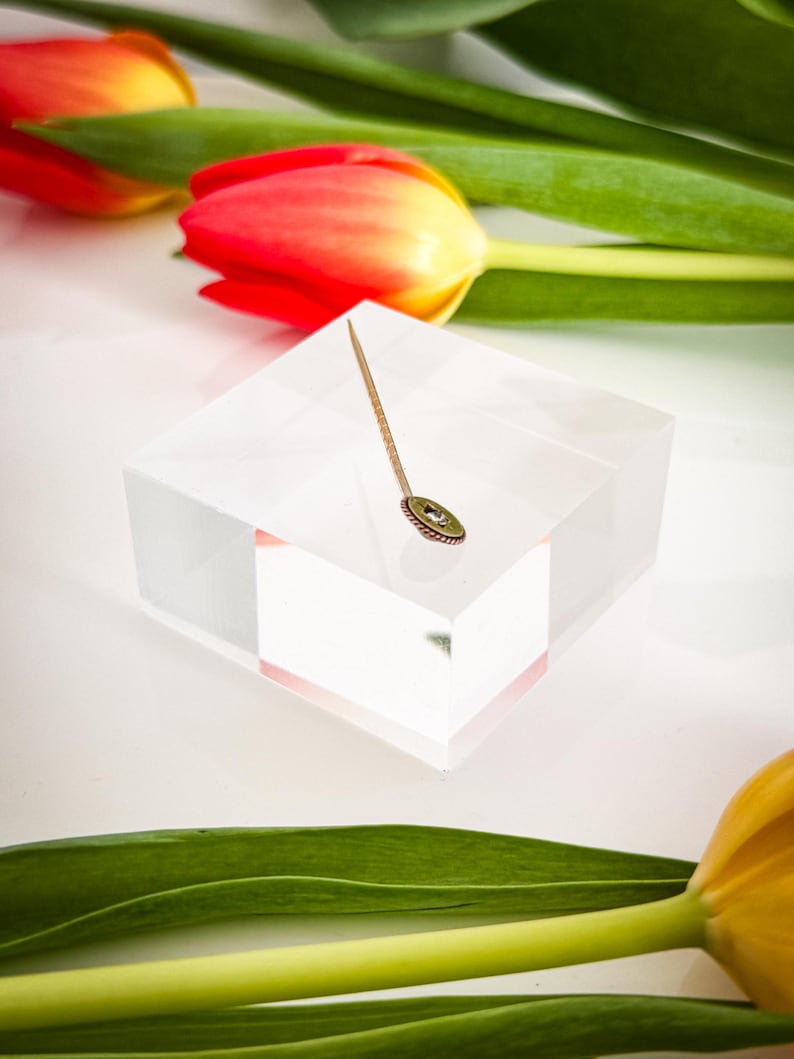 May include: A gold-toned stick pin with a small, round, green and gold decorative element. The pin is displayed on a clear, square block, with red and yellow tulips and green leaves in the background.