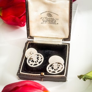 May include: Silver cufflinks with a thistle design, presented in a vintage black and brown jewelry box. The box is lined with cream-colored satin. The text on the box reads "JEWELLERS BY APPOINTMENT" and "H.L. Brown & Son Ltd."