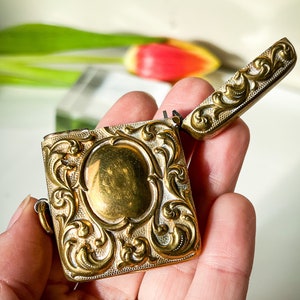 K&ouml;nnte beinhalten: Ein verziertes, goldfarbenes Scharnier-Streichholz-Sicherungsk&auml;stchen mit floralem Muster. Das Streichholz-Sicherungsk&auml;stchen ist ge&ouml;ffnet und der Deckel ist angehoben.
