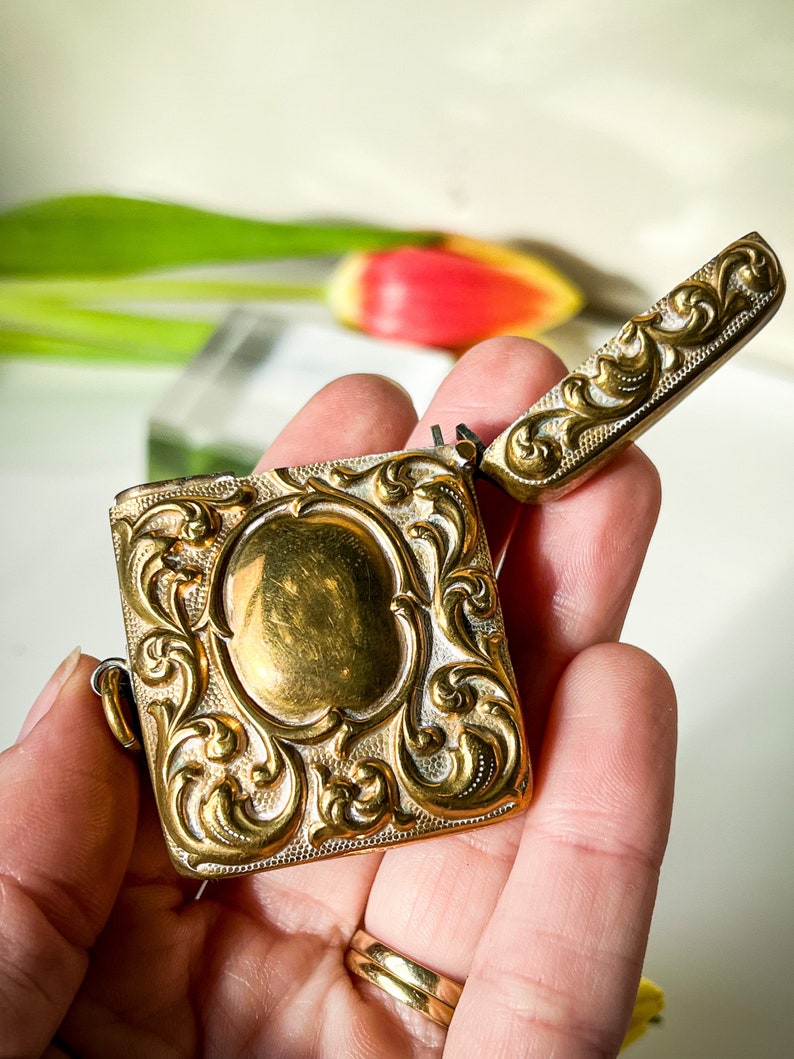 K&ouml;nnte beinhalten: Ein verziertes, goldfarbenes Scharnier-Streichholz-Sicherungsk&auml;stchen mit floralem Muster. Das Streichholz-Sicherungsk&auml;stchen ist ge&ouml;ffnet und der Deckel ist angehoben.