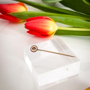 May include: A vintage-style lapel pin with a circular, light brown face and a small, diamond-shaped design in the center. The pin is set on a clear acrylic block, with red and yellow tulips in the background.