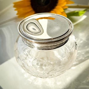 May include: A clear glass jar with a silver lid. The jar features an etched floral pattern. A yellow sunflower is in the background.