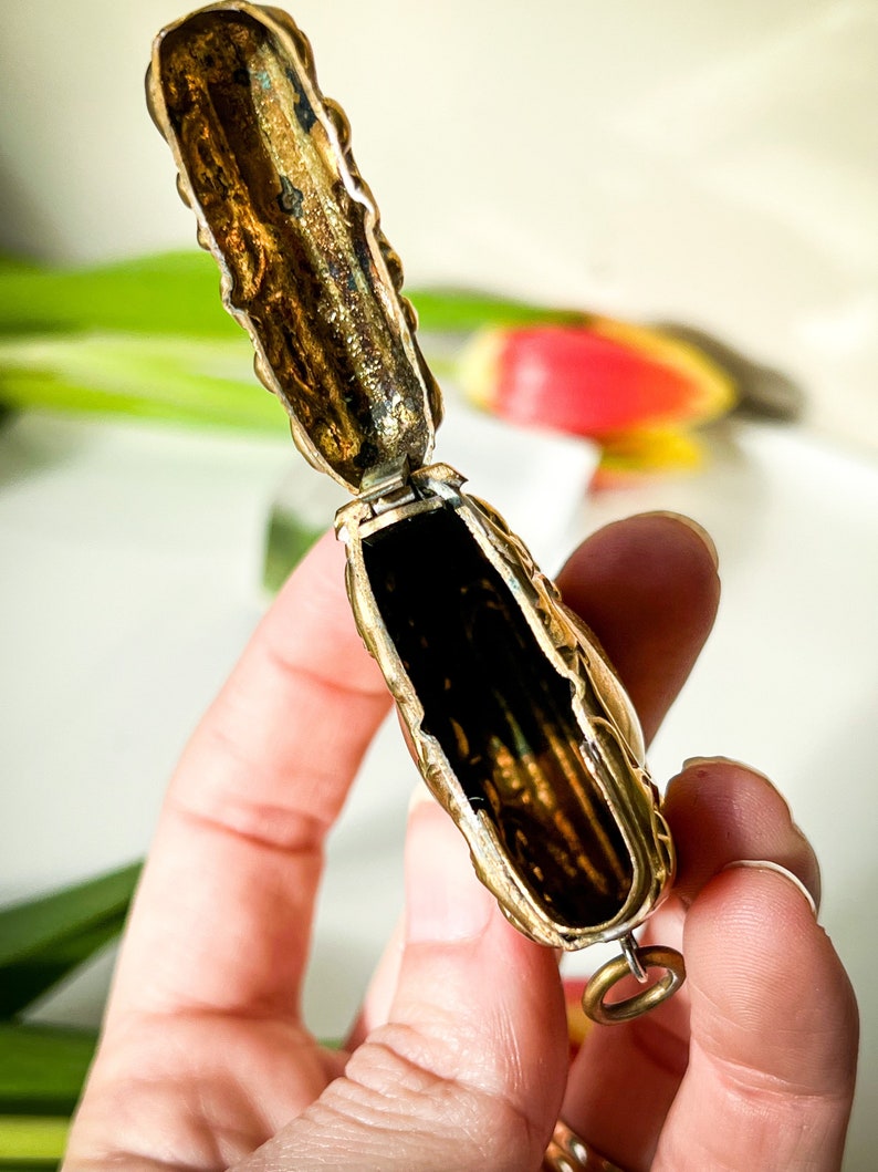K&ouml;nnte beinhalten: Ein verziertes, goldfarbenes, Scharnier-Medaillon mit einem detaillierten Blumendesign. Das Medaillon ist ge&ouml;ffnet und zeigt ein leeres Inneres. Es hat einen kleinen Ring, der an der Unterseite befestigt ist.