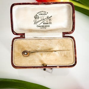 May include: An open, antique jewelry box with a velvet interior holds a gold stick pin. The pin features a small, round, diamond-like stone. The box is burgundy with the text "Goldsmiths & Jewellers" and "High St, Colchester".