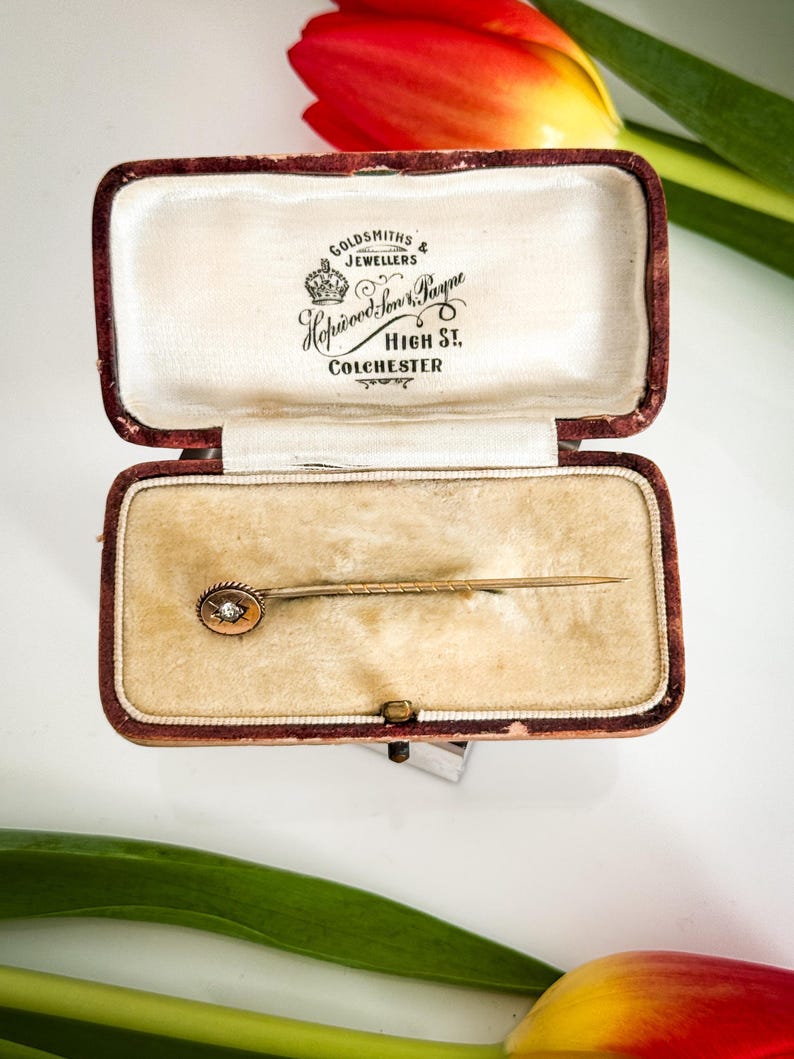 May include: An open, antique jewelry box with a velvet interior holds a gold stick pin. The pin features a small, round, diamond-like stone. The box is burgundy with the text "Goldsmiths & Jewellers" and "High St, Colchester".
