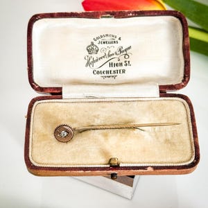 May include: An antique gold-toned stick pin with a small, round diamond set in a circular frame, displayed in a vintage maroon jewelry box with the text "Goldsmiths & Jewellers Hopwood Son & Payne High St. Colchester."
