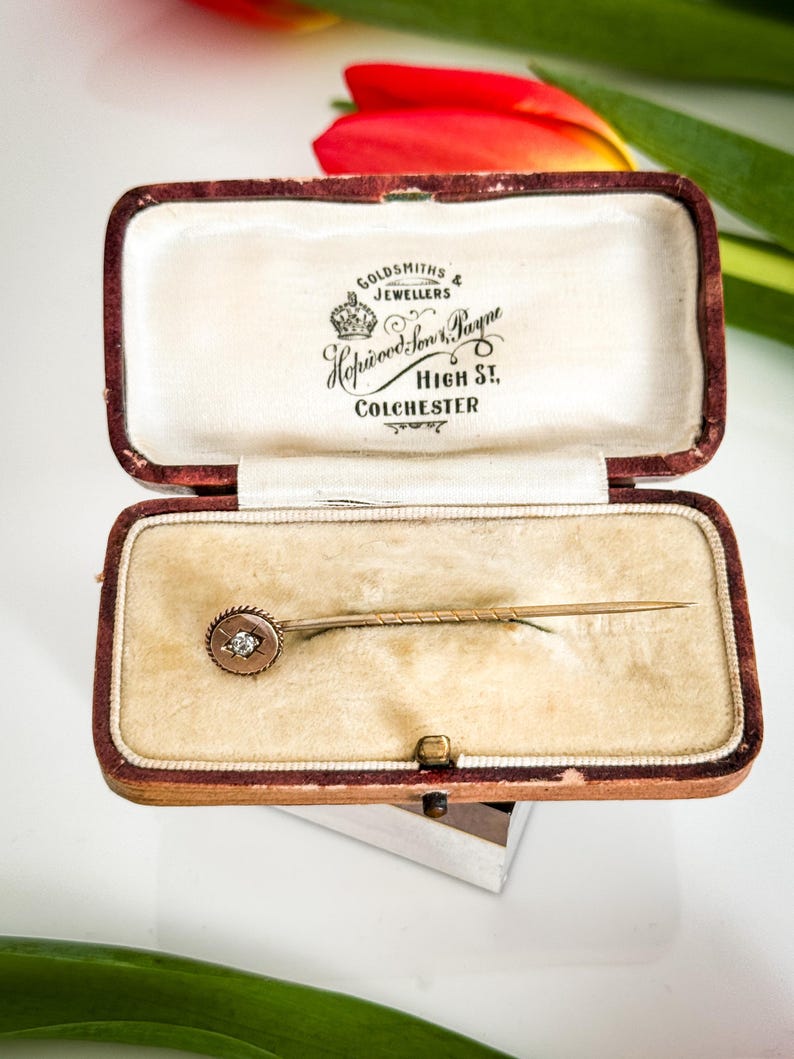 May include: An antique gold-toned stick pin with a small, round diamond set in a circular frame, displayed in a vintage maroon jewelry box with the text "Goldsmiths & Jewellers Hopwood Son & Payne High St. Colchester."